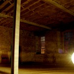 a photograph from PECo theatre's Points of Departure project. A sparkly disco ball hangs in the foreground inside an old warehouse, lighting up a thin pillar and the back wall