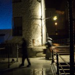 The shadowy and out of focus image of two performers moving on a landing jetty at night with the deep blue of a projection screen showing behind them