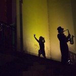 a saxophonist and a dancer are silhouetted against an grand building awash in yellow light