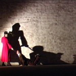 in a shadowy room, a woman in a red coat with a commanding manner stands above a man on all fours, their larger than life shadows fall on the brick wal behind them