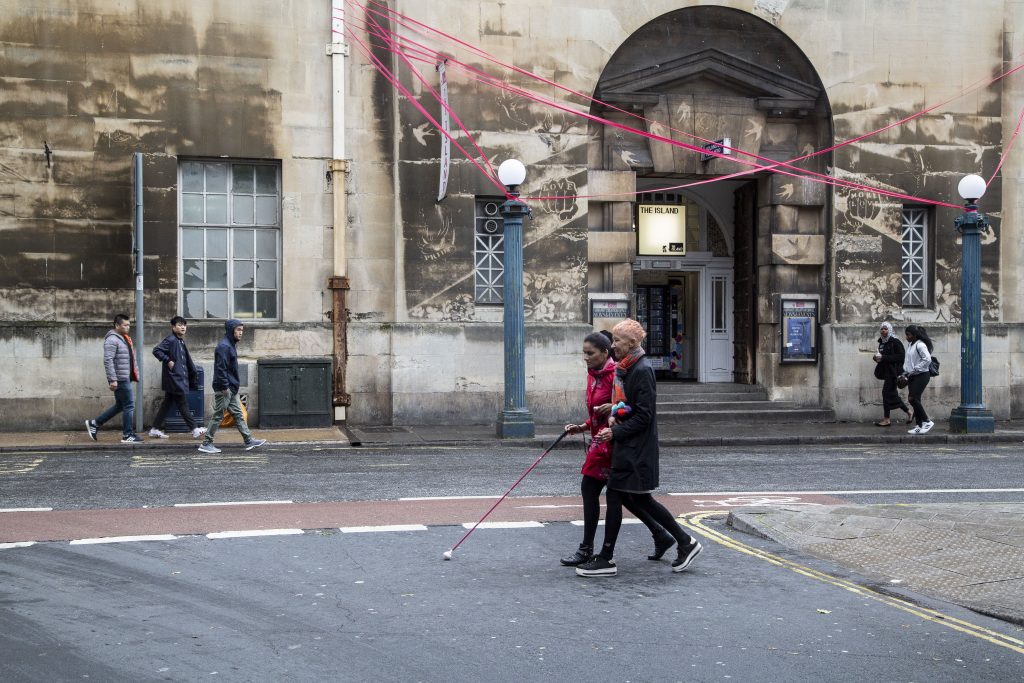two women walk arm in arm along a city street, one woman is using a long pink cane to navigate by. The pink of her cane is echoed by pink festoons hanging in the street above them. They are smiling.