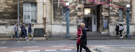 two women walk arm in arm along a city street, one woman is using a long pink cane to navigate by. The pink of her cane is echoed by pink festoons hanging in the street above them. They are smiling.