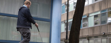 a lone journey taker from the City of Threads co-creative group navigates a street corner between a building and a street tree, using his long cane stretched out in front of him